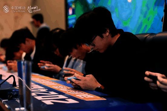 选手在比赛区专注操作手机，周围环绕着裁判与工作人员，背景为大型赛事LOGO